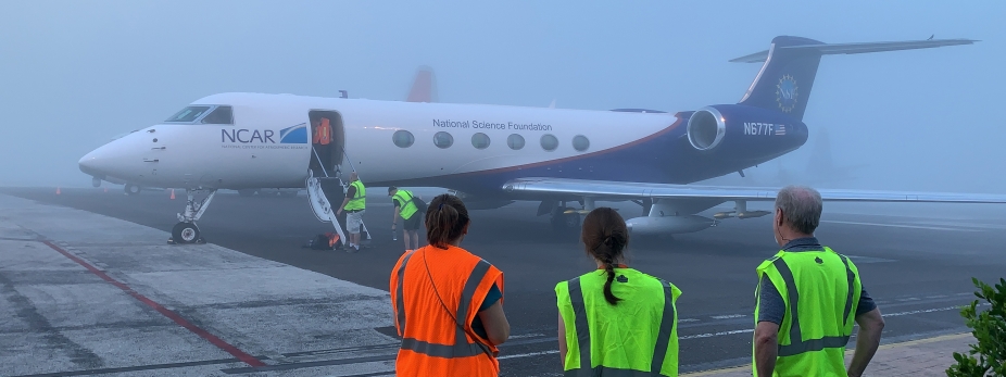 Scientists and engineers stand on the tarmac in front of a plane on a foggy morning.