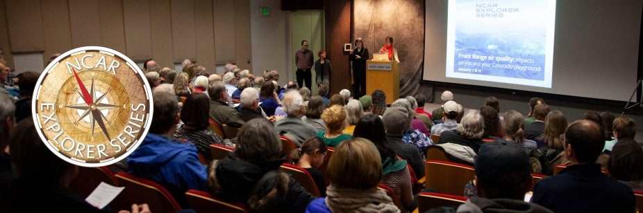 A large audience sits in an auditorium listening to a presenter on stage