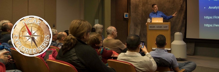 A large audience sits in an auditorium listening to a presenter on stage