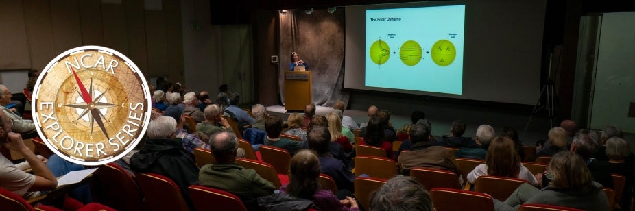 A large audience sits in an auditorium listening to a presenter on stage