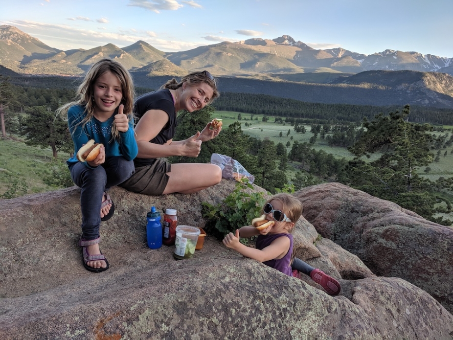 Emily Fischer eating lunch with her daughters on a rock with trees and mountains in the background.