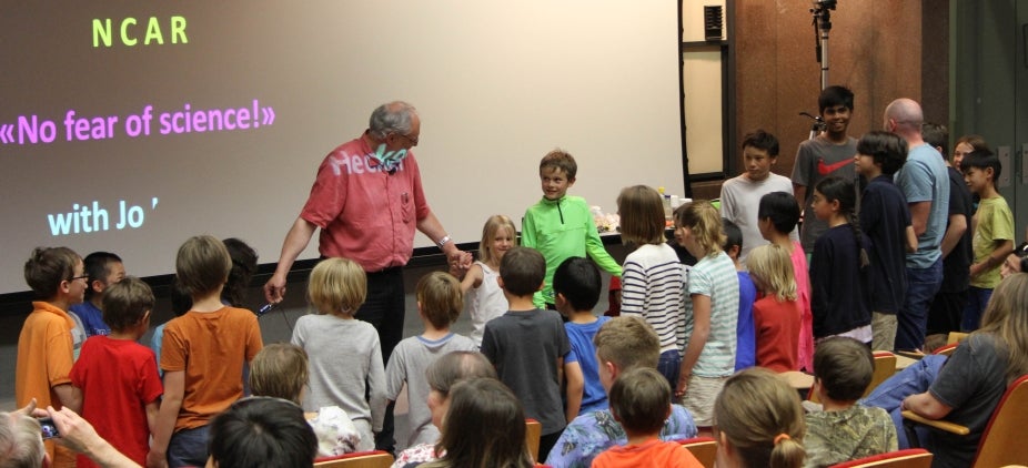 Jo Hecker and a group of kids from the audience on stage to perform a science experiment. A projection in the background says "No Fear of Science!" with Jo