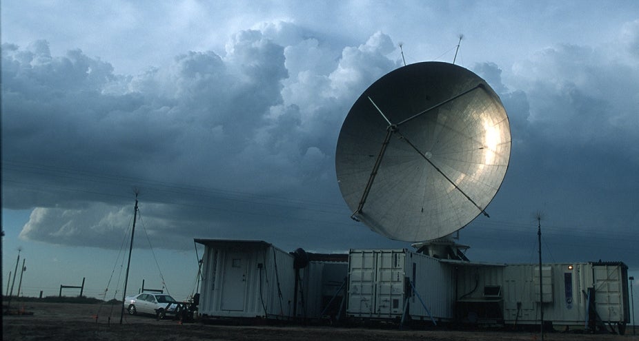 Image of a large ground radar placed above large metal containers. Clouds in the background indicate a looming storm.