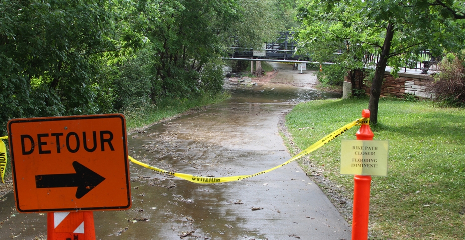 A detour sign and a caution tape block the bike path due to imminent flooding of the Boulder Creek.