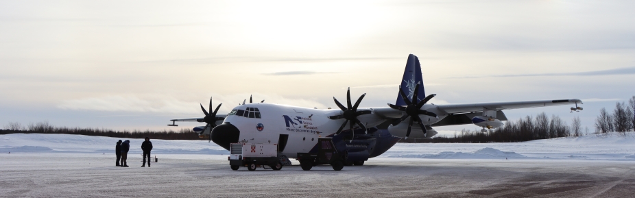 A C-130 research aircraft sits on a snowy runway, silhouetted by the sun