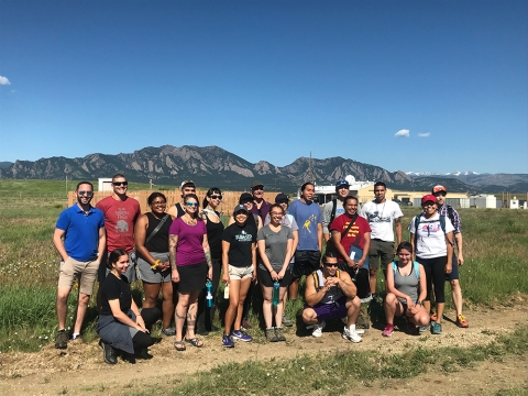 A large group of students and mentors stand in an open field, with weather instrumentation and mountains in the background