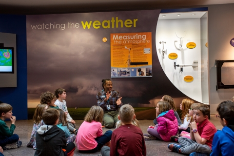 Tim Barnes kneels on the floor in front of a weather exhibit with a group of kids sitting around him