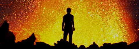 silhouette of person standing in front of a volcanic eruption. Yellow lava flares in front of him.