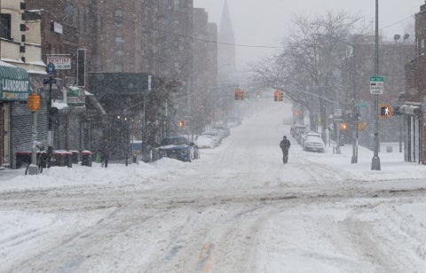 A city street covered in snow during a snowstorm, with a lone person walking in the road.