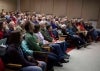 A large audience sits in an auditorium listening to a presenter